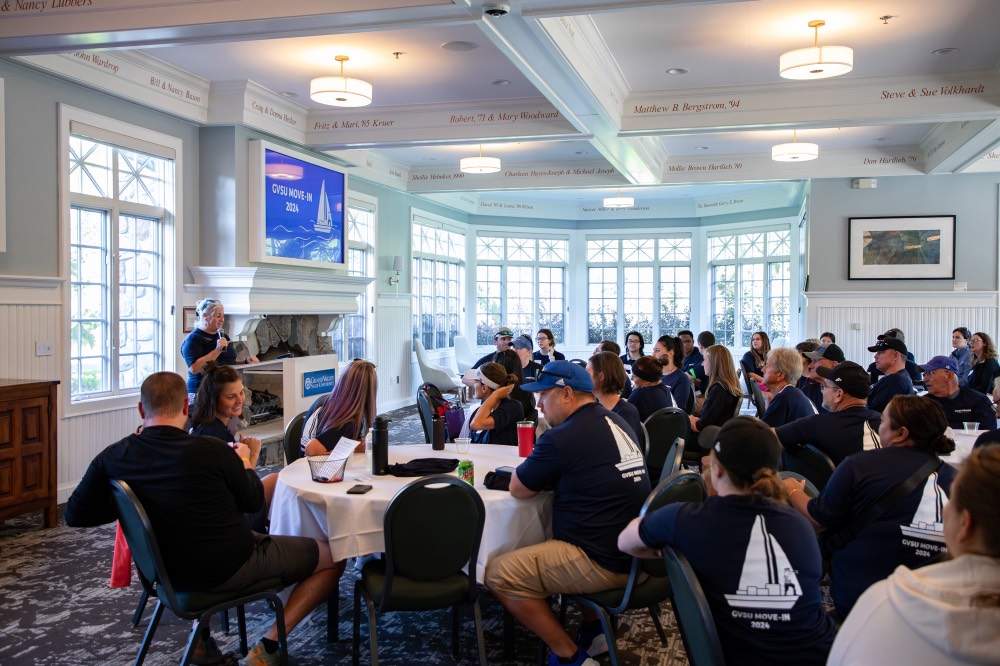 Large group of GVSU alumni volunteers sit at tables listening to Susan Proctor